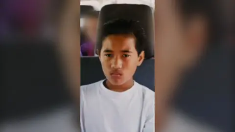 A young boy with short black hair sits in a motor vehicle. He is wearing a white T-shirt and looks upset.