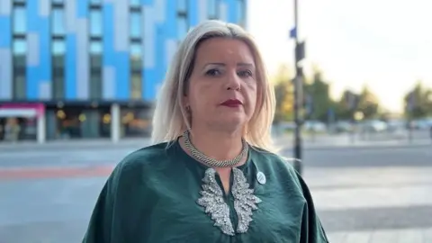 Tatiana is wearing a silver chain and a green top and is behind Slough high street. 
