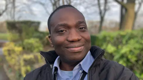 Elizabeth Baines/BBC A man smiles in woodland, he wears a blue shirt