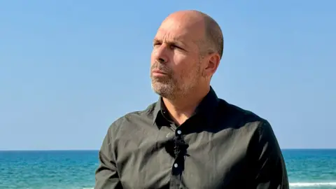 Oren Rosenfeld / BBC Roy Salomon, a man with a shaved head and stubble, with an ear on his left shirt and standing near the sea