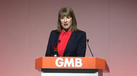 Reeves at a podium with the letters GMB on it. She is wearing a dark jacket and a red shirt.