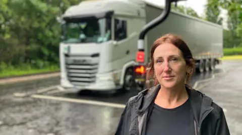 A woman in a black rainjacket is stood by a crossing on a road. There is an HGV driving past