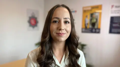 Jo Webb wearing a light coloured shirt with long brown hair smiling at the camera. She is inside a white room with posters and the background is out of focus.