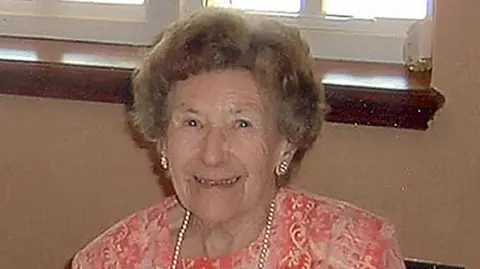 Cambridgeshire Police/PA Media Una Crown is smiling at the camera while sitting on a chair against a light beige wall. There is a window just above her. She is wearing a pink patterned dress with a long pearl necklace and shiny earrings. She has short curly hair which is mixed with shades of light brown and grey.  