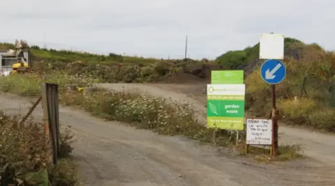 A wide shot of the entrance to the Mont Cuet landfill site. There is a dirt road and brown shrubs. There is also several signs for people visiting.