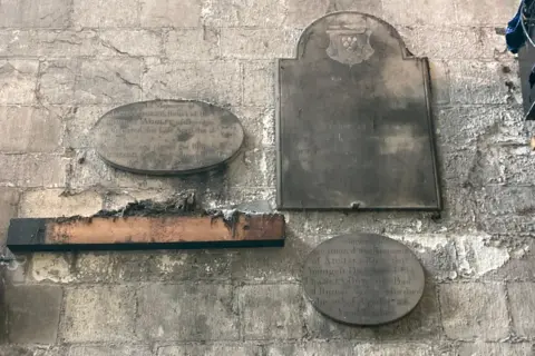 Three memorial stones and a wooden baton attached to a stone wall. Two of the memorials are oval shaped with text etched into them. The third is larger and rectangular shaped with a coat of arms at the top. The text has largely faded.