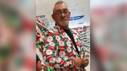 A picture of Brian Gatley. He has short hair and a short white stubbly beard and wears glasses. He wears a jacket with Santa Claus on it and is pictured in a supermarket.