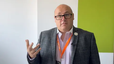 A bald man with dark glasses stands in front of a green and white background. He wears a checked suit, pink shirt and has an orange lanyard around his neck.