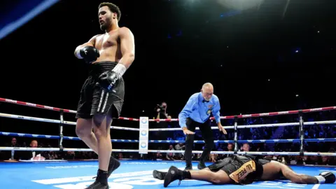 PA Media One boxer is lying face down in a boxing ring, with a referee in a blue shirt and bow tie looking down on him. Another boxer is walking away from him to the other side of the ring. 