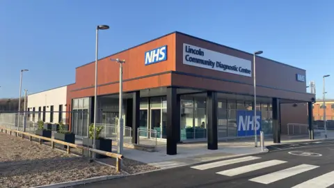 A modern building with open windows on the bottom with a brown upper floor exterior and the words NHS and Lincoln Community Diagnostic Centre written on it. 