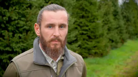 Matt Allan-Stubbs is stood front of large Christmas trees at the farm wearing a shirt and green fleece. He is looking into camera.