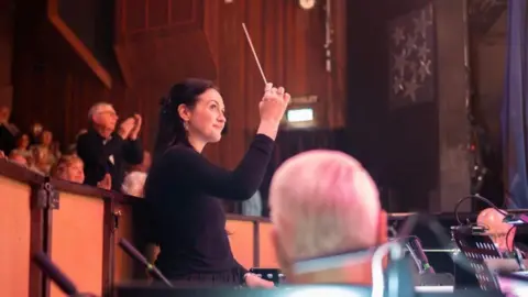 Worcestershire Acute Hospitals NHS Trust Side floor plan of a female successful achromatic conducting an orchestra. She is holding a baton and smiling. There are group clapping down her.