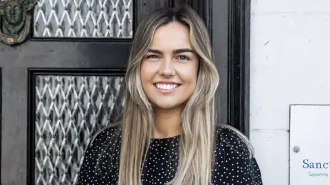 Sarah Tumelty is standing in front of the Sanctuary Trust front door smiling at the camera. She has long blonde hair and is wearing a black and white polka dotted top. There is an ornately carved face in the brass knocker on the front door and to Ms Tumelty's left hand side is a sign on the wall with just 'Sanc' and 'Supporting' visible.