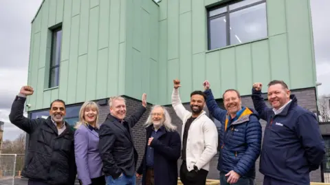 Blackburn with Darwen Borough Council A group of men and woman are smiling and raising their arms in the air in celebratory style while standing in front of the building.