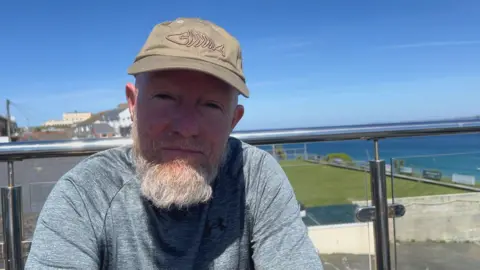 A man sitting on a balcony on a sunny day. He is looking into the camera and is wearing a cap. A shadow is over his face. Blue skies are above him and the sea is in the background. Properties are also visible behind him. 