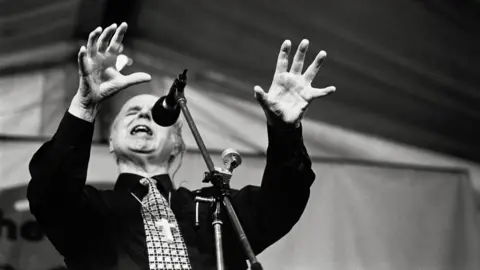 John Angerson Black and white image of Jesus Fellowship leader Noel Stanton giving a sermon with his arms raised in front of him