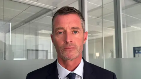 BBC Det Ch Insp Mark Hafey is wearing a dark navy suit, white shirt, and blue patterned tie stands facing the camera inside a modern office. The background features glass partition walls with frosted lower panels, metal framing, and a suspended ceiling. A coat rack with dark clothing is partially visible on the right side of the image. The scene is evenly lit with natural and indoor light, creating a professional office setting.