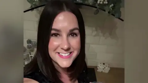 X Lucy Connolly with long straight dark hair smiling at the camera and wearing a black top. The photo is from a social media post and a table with a rock and artificial flowers is visible behind her.
