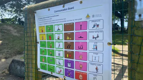 A communication board with different words and symbols on a fence. It has the words 'Communication board' at the top and the symbols are colour coded.