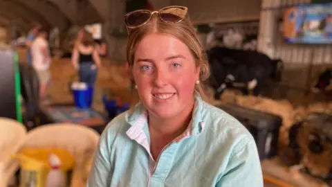 Caryl Davies, a beef and sheep farmer in one of the cattle sheds at the Royal Welsh Show. She wears a light blue jersey, and her blonde hair is tied back. A pair of sunglasses sits on the top of her head.