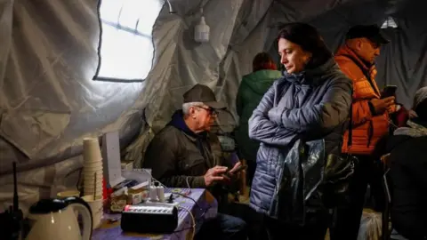Reuters A woman wearing a dark-coloured winter coat stands next to a table with cups, a kettle and plug points on it in an "invincibility centre" - where people can get an internet connection, charge their devices and warm up, amid a water and electricity supply outage in Chernihiv. A man sits on a chair behind the woman using his phone, while another is standing behind her also holding his phone. They are standing inside a large tent.