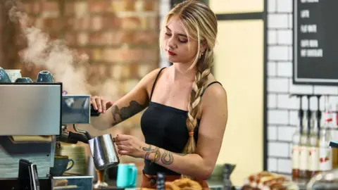 A young barista with blonde hair and tattoos serves coffee in a cafe.
