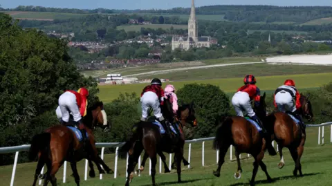 Getty Images Six horse riders head down the back straight against the backdrop of Salisbury Cathedral. They are wearing riding gear of pink and red and on brown horses.