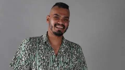 Mohaimon Mohaimon with a big smile looking at the camera, wearing a cotton printed shirt which is both striped and has flower images on it.
