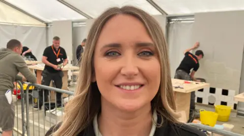 Rachel Dorovatas has long blonde highlighted hair with blue eyes and is wearing a black fleece with a light top underneath. She is standing in front of people moving wood and glass - all are wearing black t-shirts and orange lanyards.