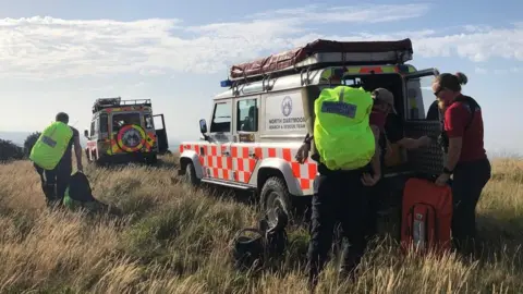 North Dartmoor Search and Rescue Team There are two Land Rovers branded North Dartmoor Search and  Rescue Team and with hi-viz emergency vehicles chevrons. They are parked in long grass. One team member is walking away wearing a large fluorescent backpack and two others are getting equipment out of the back of one of the Land Rovers. It is a sunny day.