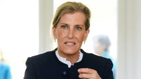 The Duchess is seated at a wooden table in a bright indoor setting, wearing a dark jacket over a light top, gesturing with both hands while speaking. The background is softly blurred, showing two other seated figures and large bright windows.