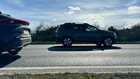 Andrew Turner/BBC Two cars pass on the Acle Straight near Great Yarmouth. The picture shows the road surface and part of the verge. It also shows clouds in a blue sky and sunshine.