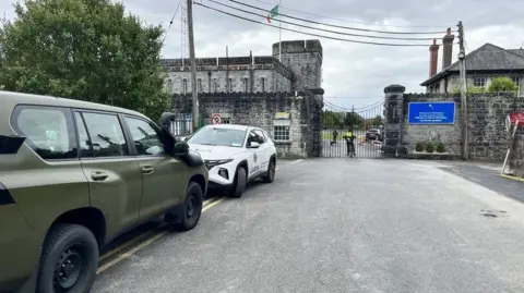 RTÉ Renmore Barracks fotografado do lado de fora do portão. O edifício é feito de pedra cinza e há um portão preto na frente dele. Uma viatura da polícia está estacionada do lado de fora.