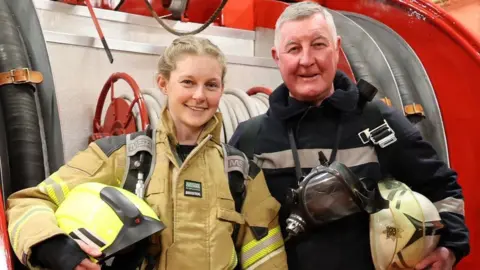 Paul Horton Media and Photography Lou Needham in the fire kit she will be wearing for the climb, with her father David in the 1986 uniform. They are both holding their helmets