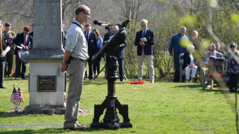 A man stands by a plinth at Markshall, with a microphone surrounded by a crowd commemorating an event.  Some of the crowd are standing, some are sitting down.  The event is held on grass with a stone monument present. Small American and British flags can be seen, sticking out from the ground.