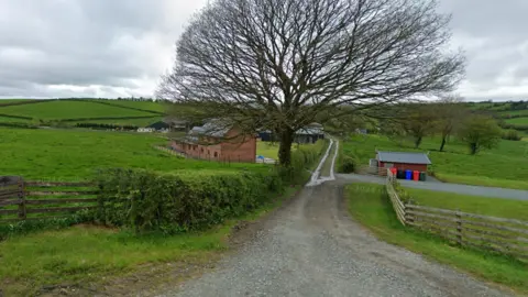 Google Tan y Dderwen Farm in Betws-yn-Rhos. A house can be seen in the centre behind a large oak tree. A lane road runs in the centre, with green fields either side. Rolling hills can be seen behind the house along with farm sheds.