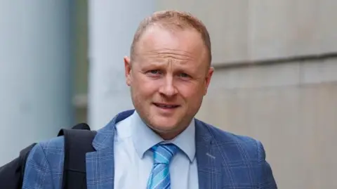 Jamie Bryson walking outside of court wearing a light blue check suit, lighter blue shirt and striped blue tie. He has a black back pack strap over his right shoulder. 
