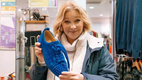 BBC/One Tribe TV Joanna Page is holding a blue shoe and looking at the camera. She is stood in a charity shop wearing a coat.