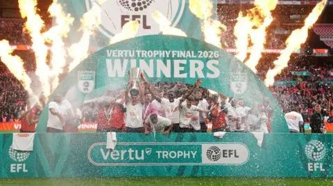 PA Media Luton Town players celebrate with the trophy after winning the Vertu Trophy final at Wembley Stadium, London. There are supporters behind them in the stands. The image is blurred due to confetti cannons going off. The players are in front of a sign and one is holding up the trophy. 