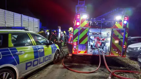 A fire engine and a police car parked at the scene of a fire in the early hours of the morning. The fire engine has various lights on its body switched on while firefighters stand next to it wearing various fire fighting apparatus. 