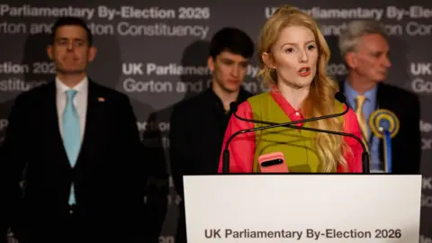 Hannah Spencer looks to the right of frame as she speaks while standing at a lecturn that reads 'UK parliamentary by-election 2026; Gorton and Denton Constituency', with three other candidates standing onstage behind her.