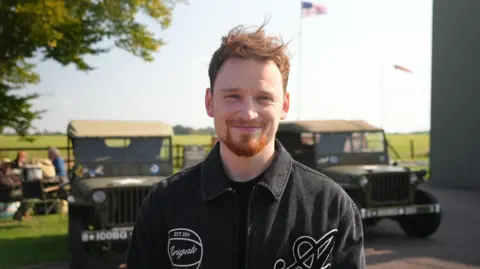 Shaun Whitmore/BBC Louis Greatorex wearing a black shirt and standing in front of two military vehicles and an American flag in the distance