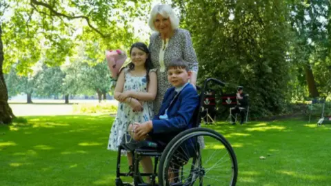 PA Media Tony Hudgell with Queen Camilla and Lyla O'Donovan. They have been pictured outside in the sun on the grass. Tony is in a wheel chair - he is an amputee missing both legs. He wears a suit. Lyla has long dark hair and wears a summer dress. Queen Camilla wears a floral dress and is holding a bouquet of flowers. There is a brass band in the background and trees. 