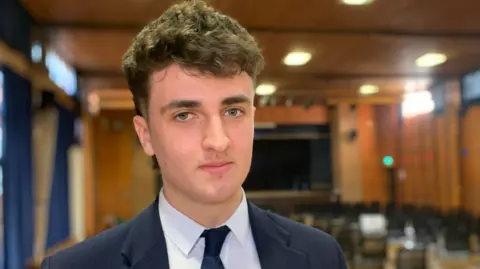 Taylor, who is wearing a navy school uniform, has thick brown hair and blue eyes, looks into the camera with a neutral expression