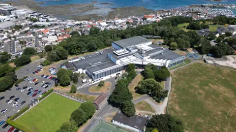 BBC An aerial photograph showing a large building complex set within landscaped grounds. Surrounding the building are car parks filled with vehicles, a green bowling lawn, open playing fields, and a children's playground. Beyond the site lies a densely built coastal town, a marina with moored boats, and a rocky shoreline exposed by low tide, with blue sea stretching across the top of the image.