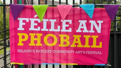 A pink poster with white and yellow writing which says Féile an Phobail on it against black railings