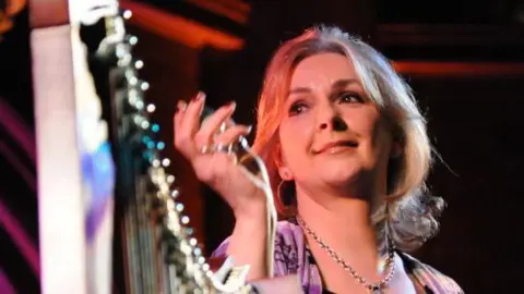 Getty Images Moya Brennan is standing beside a harp. She is wearing a silver necklace and has long blonde hair. Her right hand is raised.