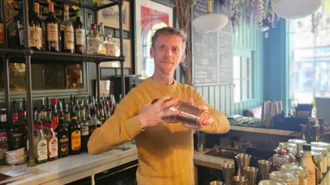 A blond man in a yellow jumper is holding a cocktail shaker. He is standing inside a bar.
