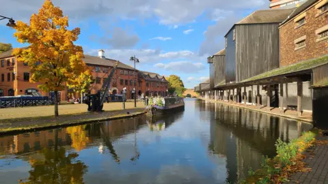 BBC Weather Watchers/pete the pilot A canal basin with restored and rebuilt industrial buildings around it and a boat moored in the centre of the picture. A tree with yellowing leaves overlooks the water beneath a blue sky punctuated by grey clouds. 