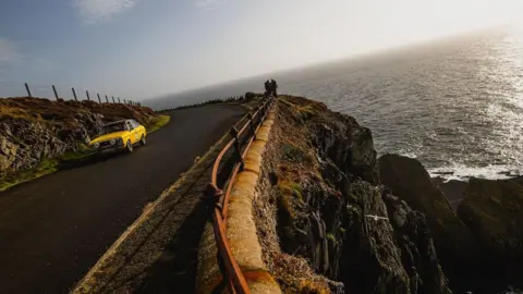A classic yellow car drives round a corner at Marine Drive on a clear day.
